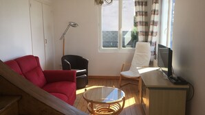 Una habitación con un sofá rojo, una silla negra, una silla blanca, una mesa de vidrio, un escritorio de madera, un televisor y una ventana con cortinas.