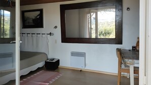 Een slaapkamer met een bed, een spiegel, een radiator, een klein tafeltje met een fles en een raam met uitzicht op groen.