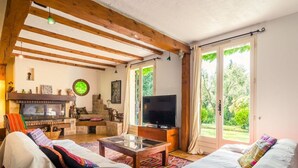 A cozy living room with a fireplace, a television, and a large window offering a view of greenery. - house / villa - 8 rooms - 8/10 persons (Auriol)