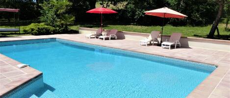 Ein rechteckiger Pool mit gefliesten Rändern, umgeben von einer gefliesten Liegefläche. Zwei rote Sonnenschirme und mehrere weiße Liegestühle sind vorhanden.