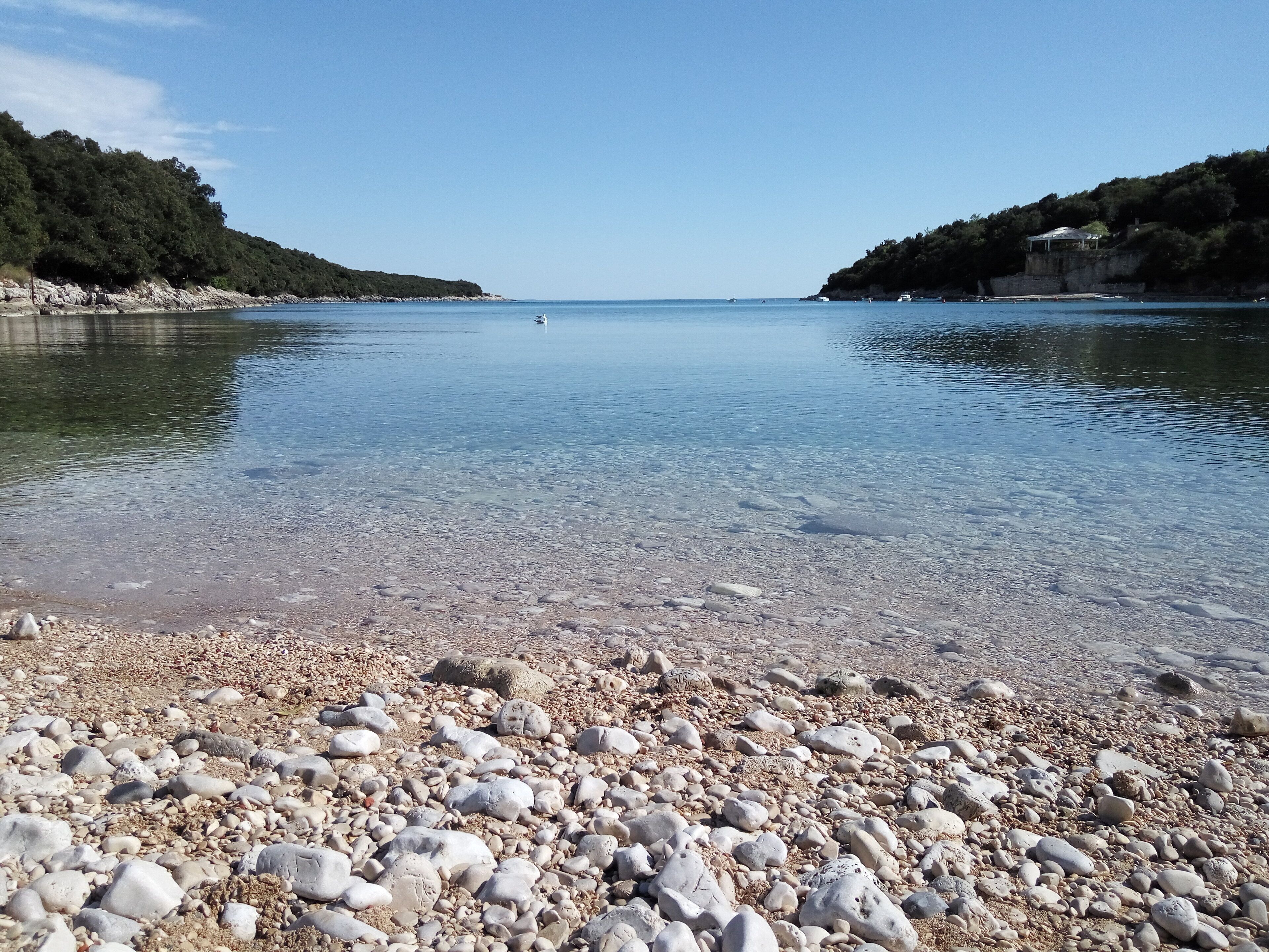 Une plage de galets, avec une eau claire et des bateaux visibles au loin.