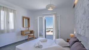 Una camera d'albergo con un letto, una scrivania, una sedia, uno specchio, una porta che dà su un balcone e vista sul mare.