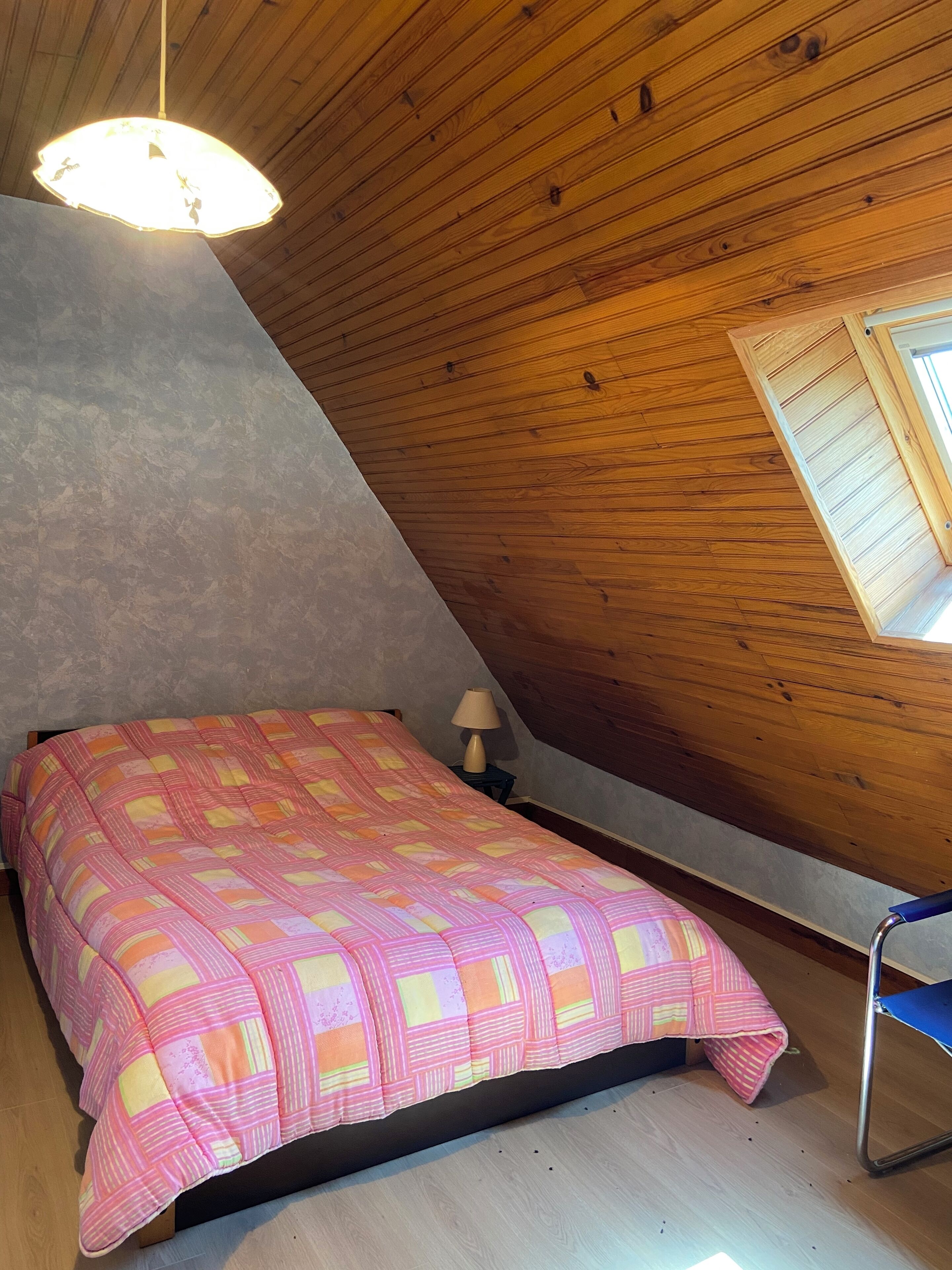 Une chambre avec un plafond en pente, un lit recouvert d’une couverture aux couleurs vives, une lampe de chevet et une fenêtre avec des volets.