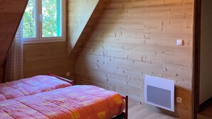Quarto com teto inclinado, cama com colcha rosa e laranja, luminária no teto e janela com vista para árvores.