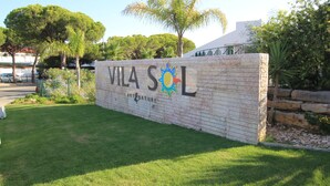 Skilt for Vila Sol med et logo af sol og natur, omgivet af grønt og en klar himmel.