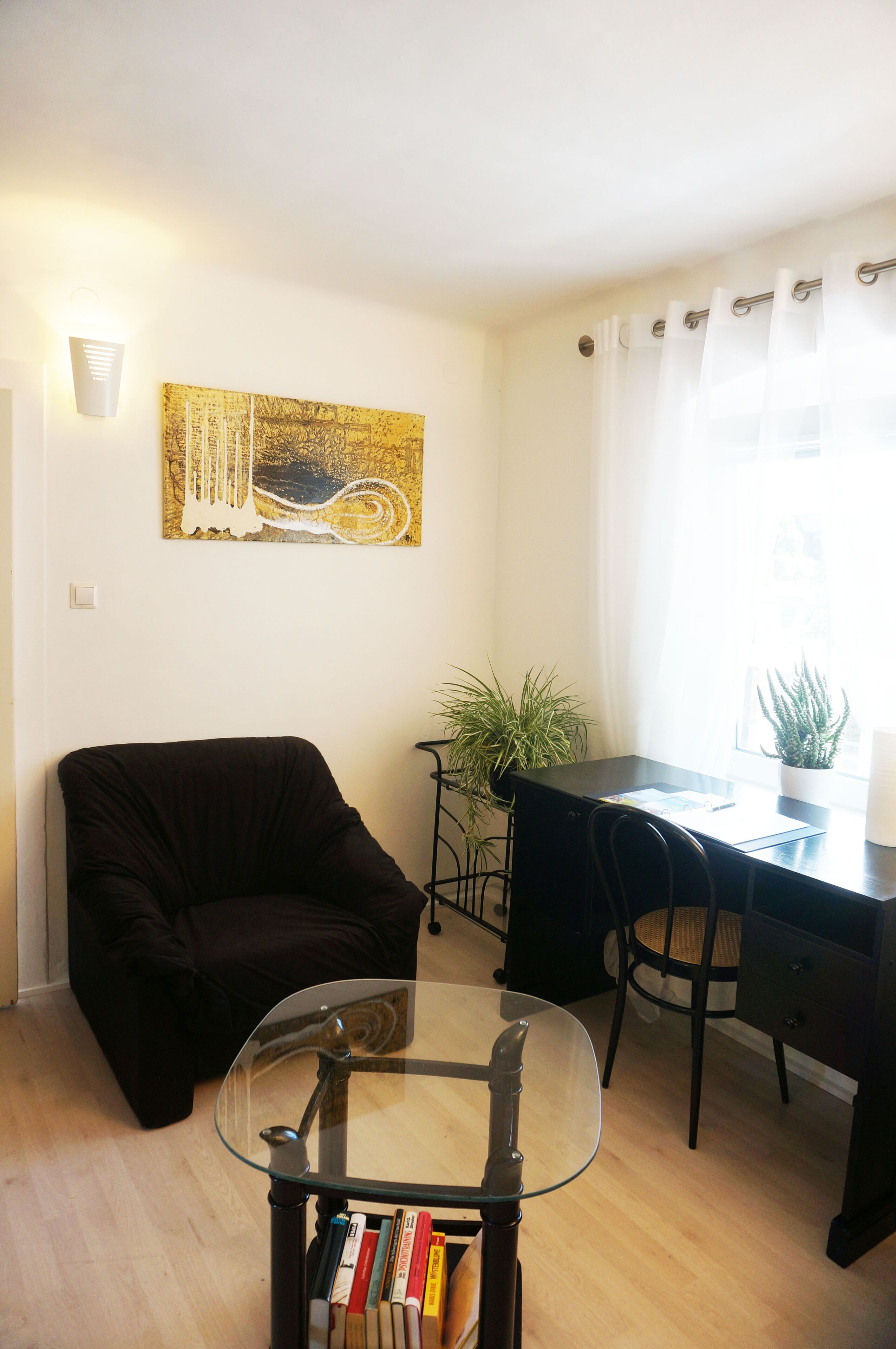A living room with a glass-top coffee table, a black sofa, a desk with a chair, a potted plant, and a painting on the wall.