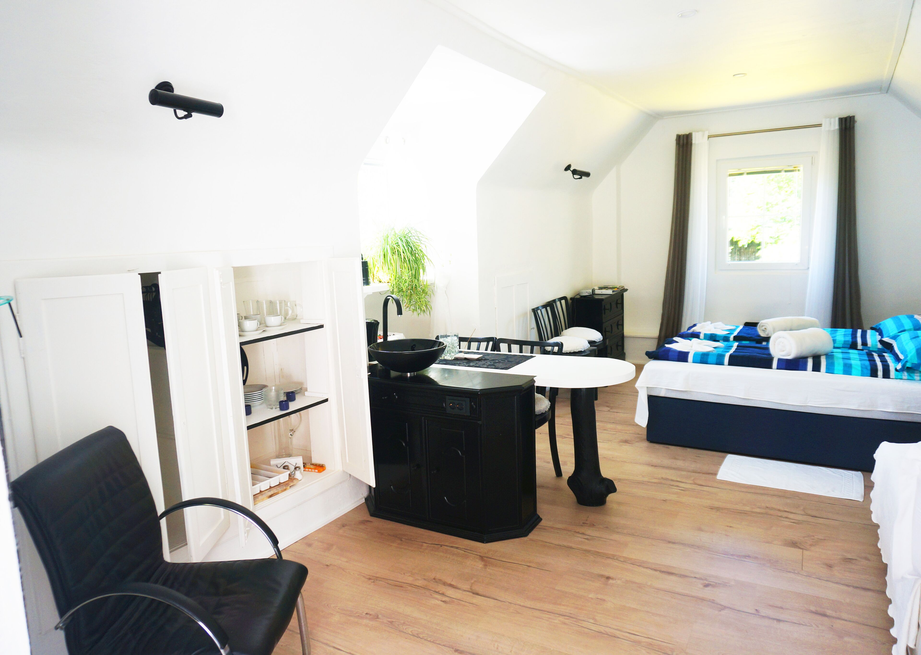 A compact hotel room with a kitchenette, a dining area, a bed with blue bedding, and a large window with curtains.