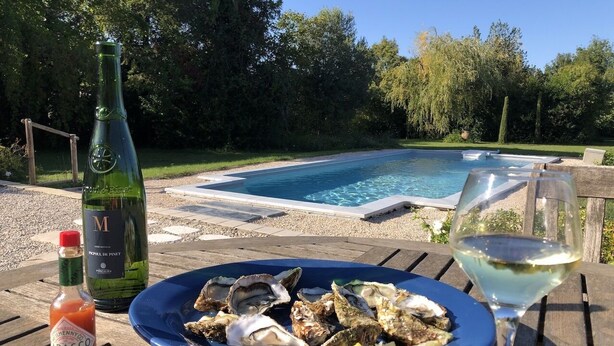 Une table à manger avec une assiette d’huîtres, une bouteille de sauce piquante et un verre de vin blanc, le tout installé à l’extérieur, près d’une piscine.