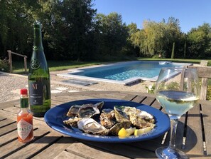 A dining table with a plate of oysters, a bottle of hot sauce, and a glass of white wine, set outdoors near a swimming pool. - La Maison Blanche in the countryside with large private garden, terrace and pool (Saint-Rémy-De-Provence)