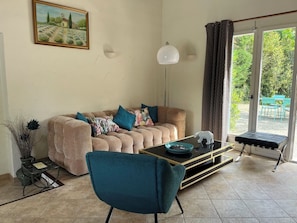 Un salon comprenant un canapé, une table basse, un fauteuil, un tableau, une lampe et une vue à travers une porte vitrée.