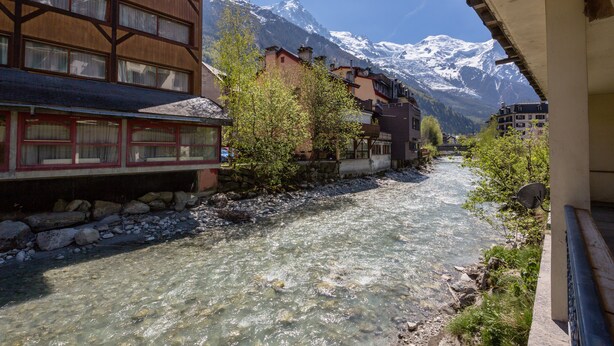 A river flowing through a town with buildings on either side and snow-capped mountains in the background. - Chamonix centre 2BR - Fireplace, stone walls, balcony with View of Mont Blanc (Chamonix-Mont-Blanc)