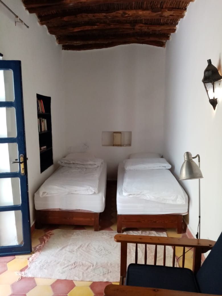 Une petite pièce avec deux lits simples, un plafond en bois, une porte, une étagère fixée au mur et un lampadaire.