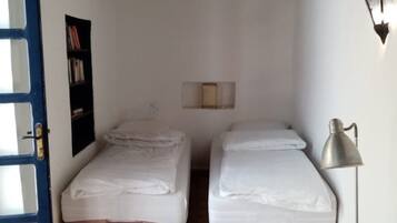 Une petite pièce avec deux lits simples, un plafond en bois, une porte, une étagère fixée au mur et un lampadaire.