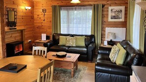A cozy cabin interior with a fireplace, leather sofas, a wooden dining table, and a large window with curtains. - Cherry Lodge, Cedar Springs, Self Catering Log Cabin, Heacham,Close to the Beach (King's Lynn)