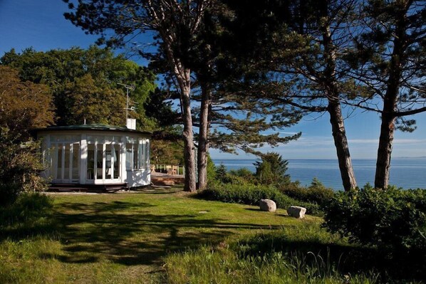 Uma cabana pequena e branca com telhado verde, cercada por árvores e vista para o mar.
