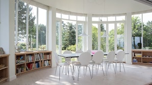 Ein heller, sonnendurchfluteter Raum mit einem großen Tisch und Stühlen, umgeben von Bücherregalen und großen Fenstern mit Blick auf Grünflächen.