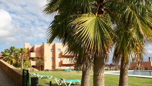 Un hotel con piscina, palmeras y un área cercada con sillones de descanso.