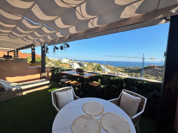 Un patio al aire libre con vistas al mar y grúas de construcción, que presenta una mesa redonda con individuales y sillas de mimbre.