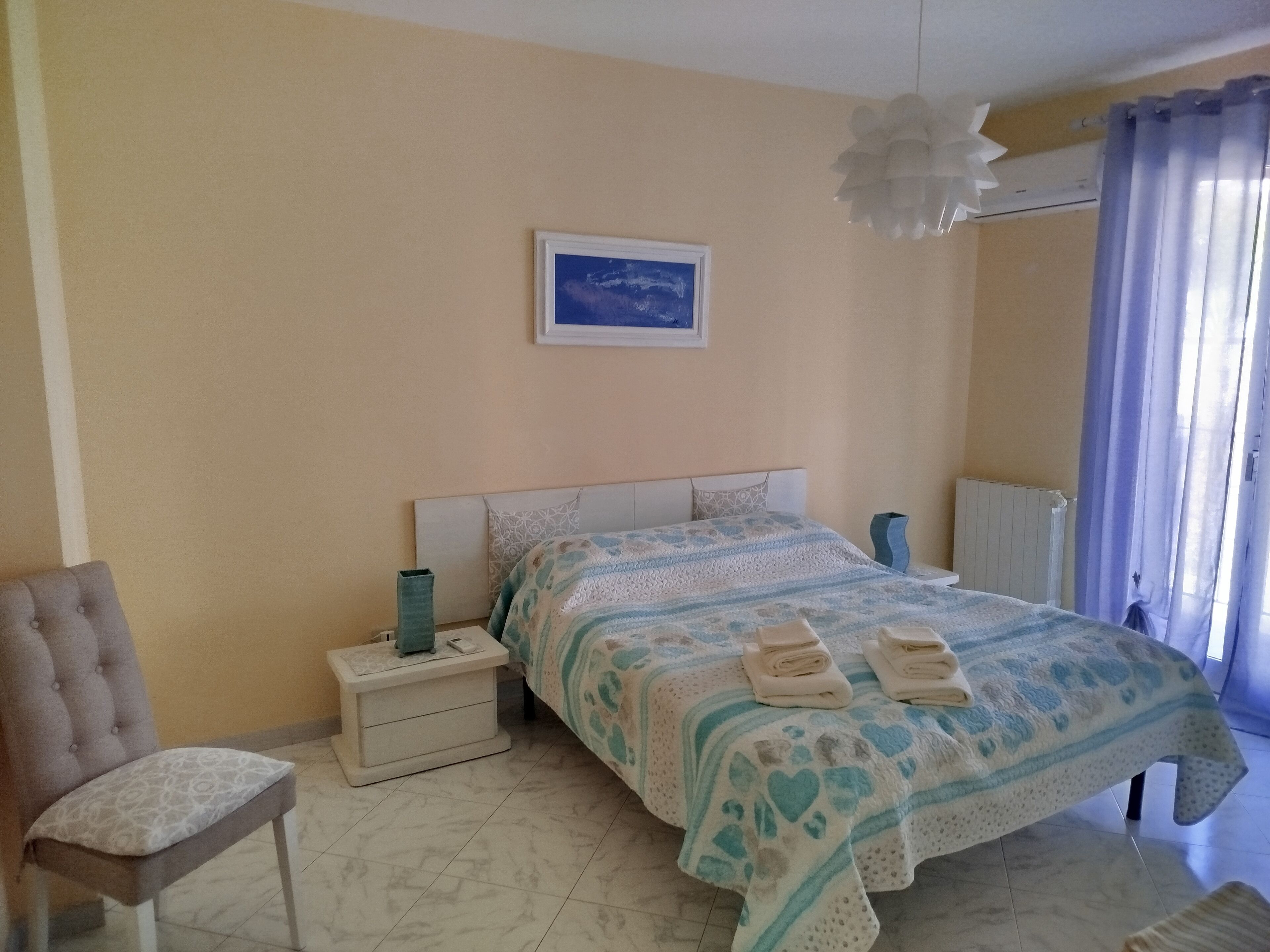 Un dormitorio con cama, mesita de noche, silla y ventana con cortinas.