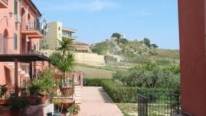 Un chemin pavé mène à un bâtiment doté d’un balcon, qui est entouré de plantes en pot et offre une vue sur une colline.