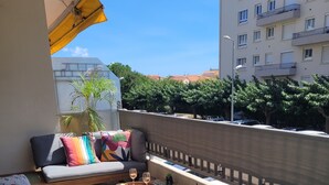 En balkon med en sofa, et bord med drikkevarer, plantekrukker og udsigt til bygninger og træer.