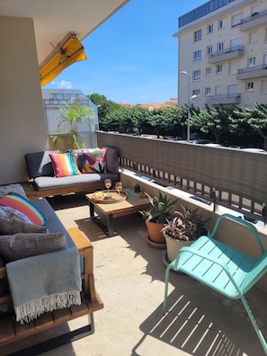 A balcony with a sofa, a table with drinks, potted plants, and a view of buildings and trees. - Large bright apartment - beside park. (Perpignan)