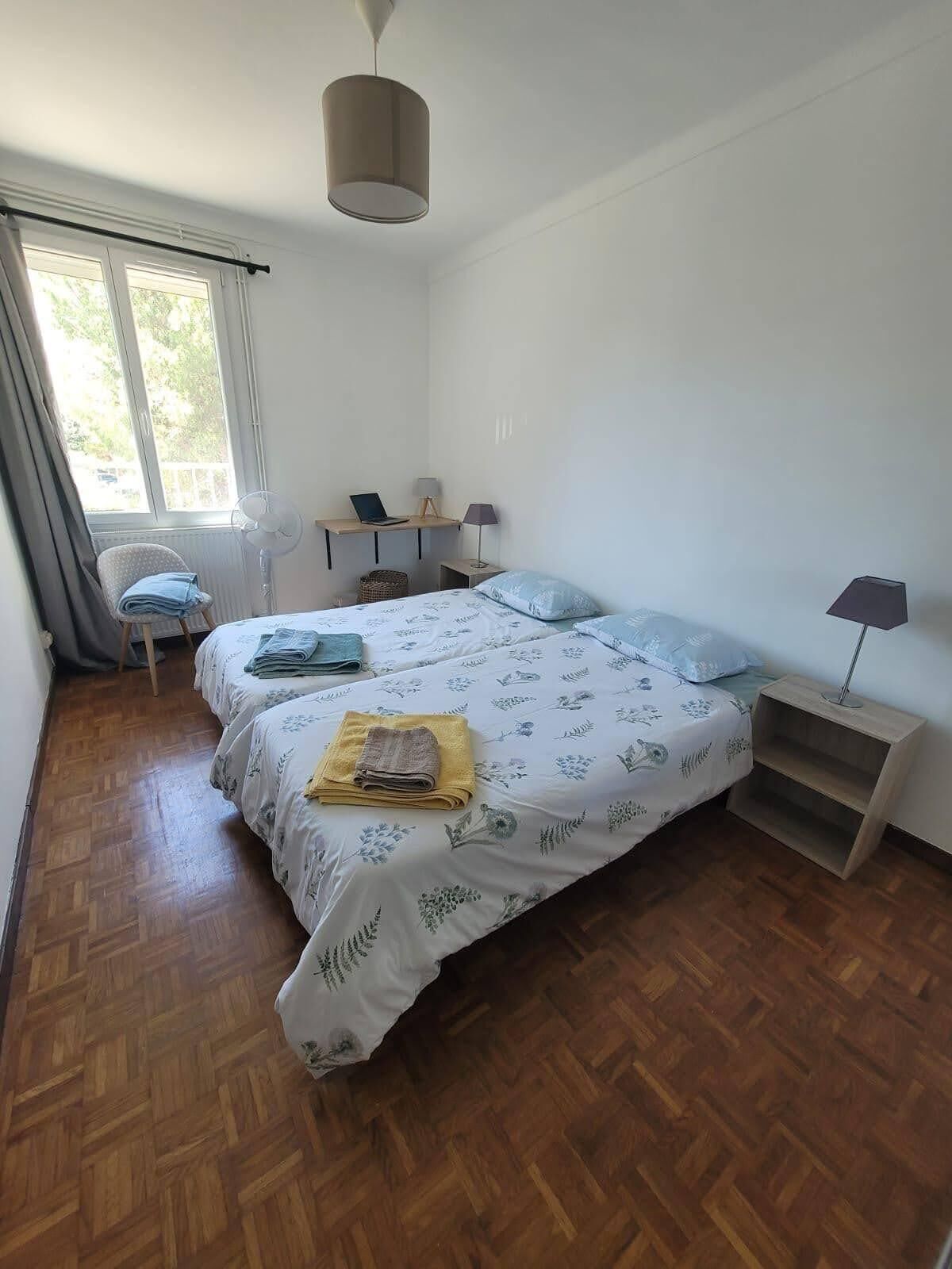 Une chambre avec deux lits, un bureau, une chaise, un ventilateur sur pied et une fenêtre avec des rideaux.