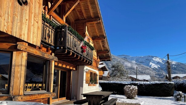 Een houten chalet met een bubbelbad op de voorgrond, een balkon met rode decoraties en een besneeuwde berg op de achtergrond.