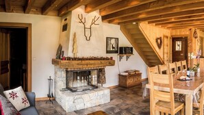 Een gezellige woonkamer met een stenen openhaard, houten balken, een eethoek met een grote tafel en stoelen, en een trap naar een ander niveau.