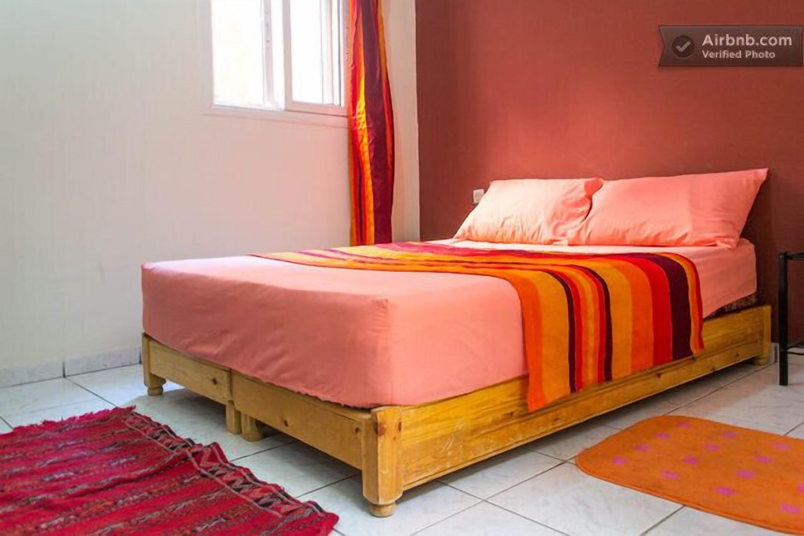 A single bed with a pink bedspread and orange and red striped blanket in a room with a window and a red curtain.