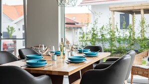 Una mesa de comedor dispuesta con platos azules y copas de vino, rodeada de sillas negras, con vista a un jardín y edificios al fondo.