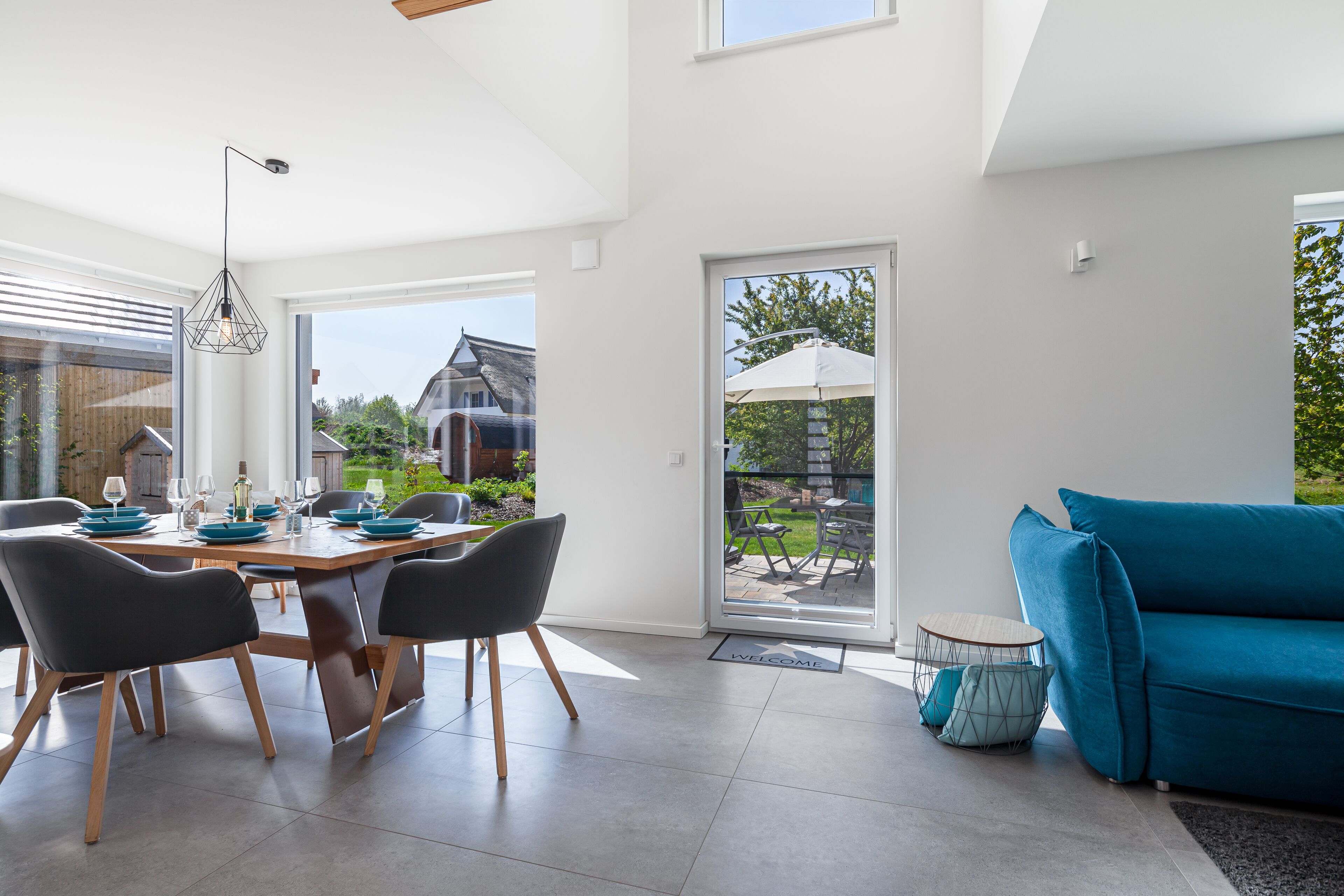 Un espace repas moderne avec une table en bois et des chaises noires, un canapé bleu et une vue sur l’extérieur grâce à de grandes fenêtres.
