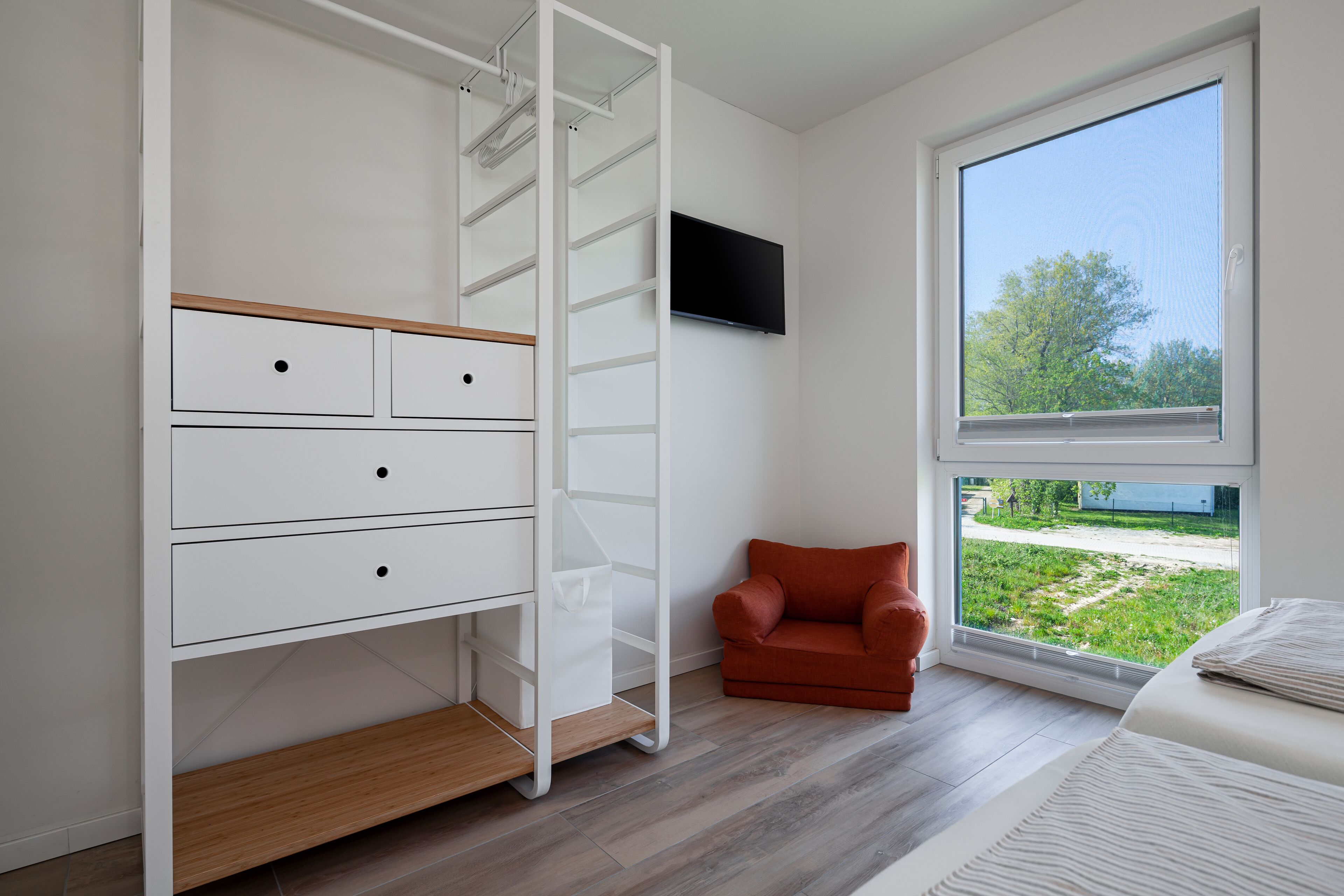 A bedroom with a bed, a TV mounted on the wall, a window with a view of greenery, and a white wardrobe with drawers and shelves.
