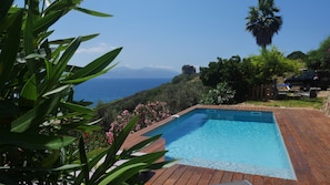 Une piscine rectangulaire avec une plage en bois, entourée d’une végétation luxuriante et offrant une vue sur la mer.
