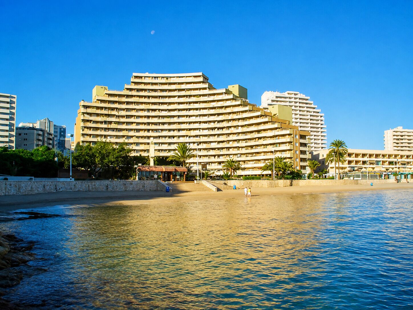 Un gran hotel con varios pisos y balcones, ubicado junto a una playa de aguas cristalinas.