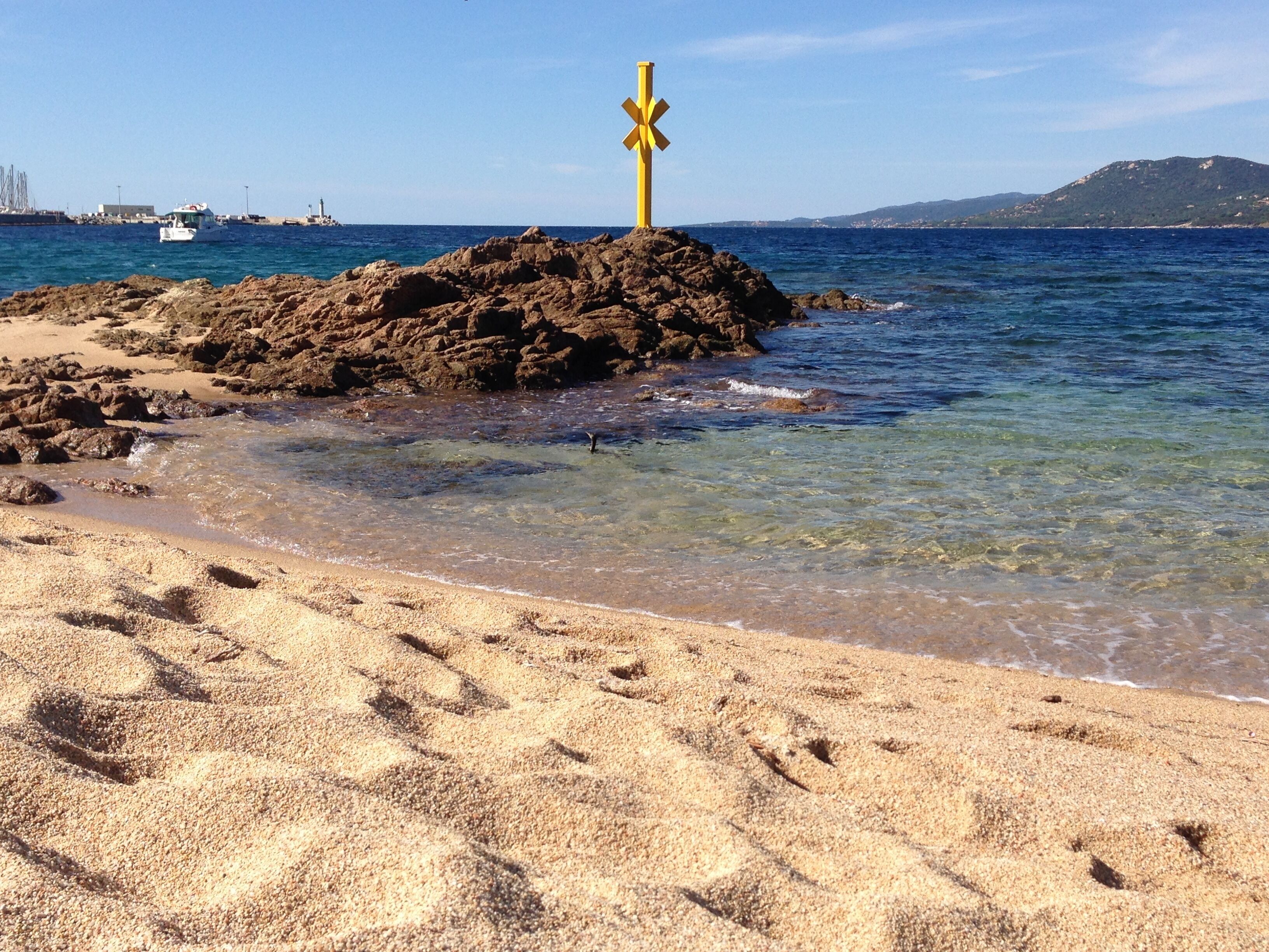 A sandy beach with footprints leading to a rocky outcrop where a yellow cross is placed.