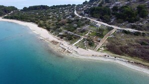 Ett flygbildsbild av ett kustområde med en strand, klart blått vatten och ett kuperat landskap med ruiner.