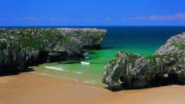 Une plage aux eaux turquoise et limpides, bordée de formations rocheuses et d’un rivage sablonneux.