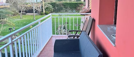 A balcony with a wicker sofa, a white railing, and a view of a green landscape with houses.