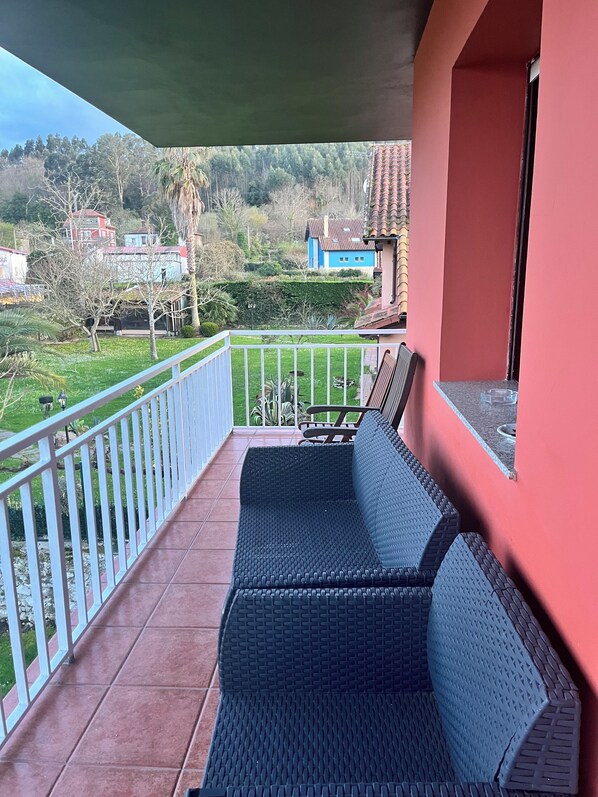 Un balcón con un sofá de mimbre, barandal blanco y vistas a un paisaje verde con casas.