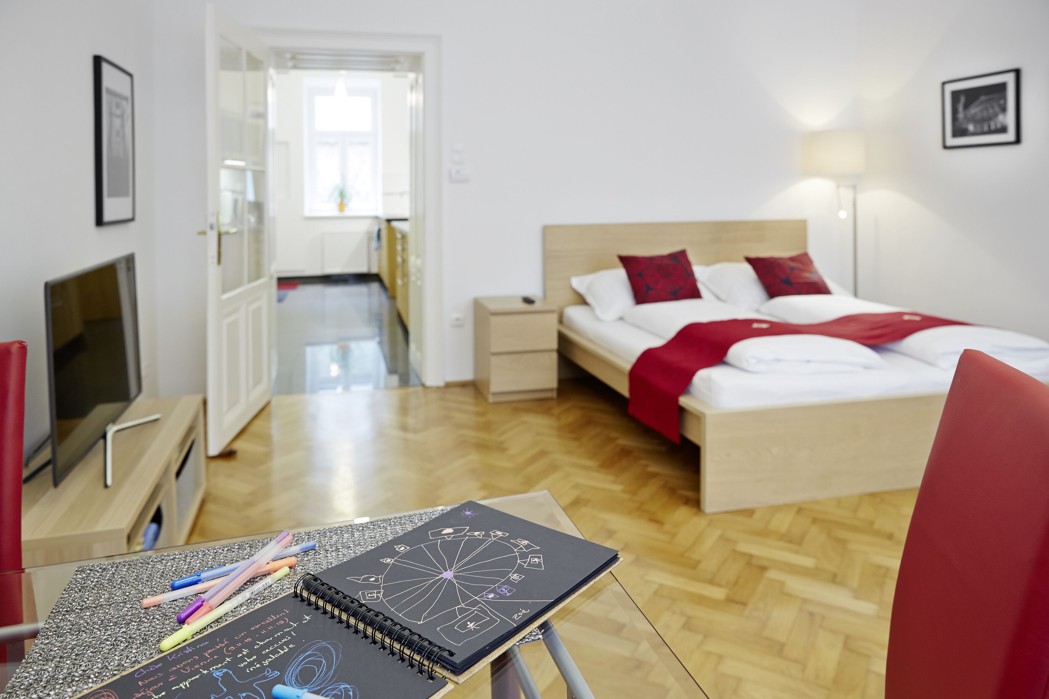 A modern hotel room with a wooden floor, a bed with red bedding, a desk with a notebook and pens, and a TV on a stand.