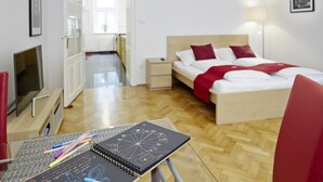 Ein modernes Hotelzimmer mit Holzboden, einem Bett mit roter Bettwäsche, einem Schreibtisch mit Notizbuch und Stiften sowie einem Fernseher auf einem Standfuß.