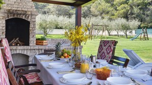 An outdoor dining area with a stone fireplace, a table set for a meal, and a view of a grassy yard with trees. - Beautiful **** villa with heated pool, summer kitchen, WiFi, grill, fitness, 1.5 km from the sea (Pula)
