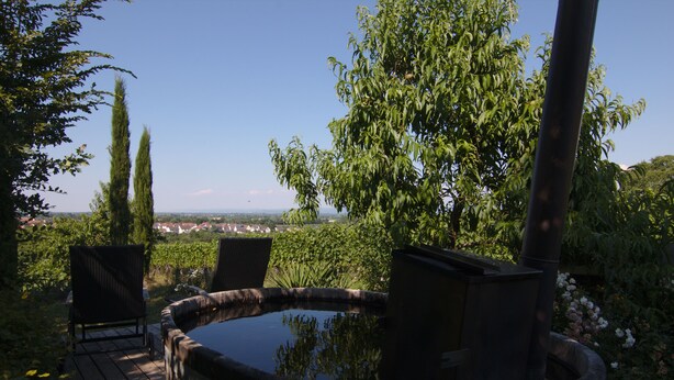 A hot tub with a wooden deck and chairs, surrounded by greenery and a view of a distant town. - Junior Suite Girardi Mountain View (Sasbach am Kaiserstuhl)
