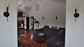 A modern living room with a gray sectional sofa, a glass coffee table, and a wooden floor. There are two wall-mounted clocks and a kitchen area visible in the background. - Junior Suite Girardi Mountain View (Sasbach am Kaiserstuhl)