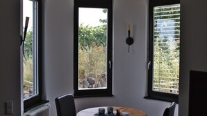 A dining area with a round table, four chairs, and three windows with blinds. - Junior Suite Girardi Mountain View (Sasbach am Kaiserstuhl)