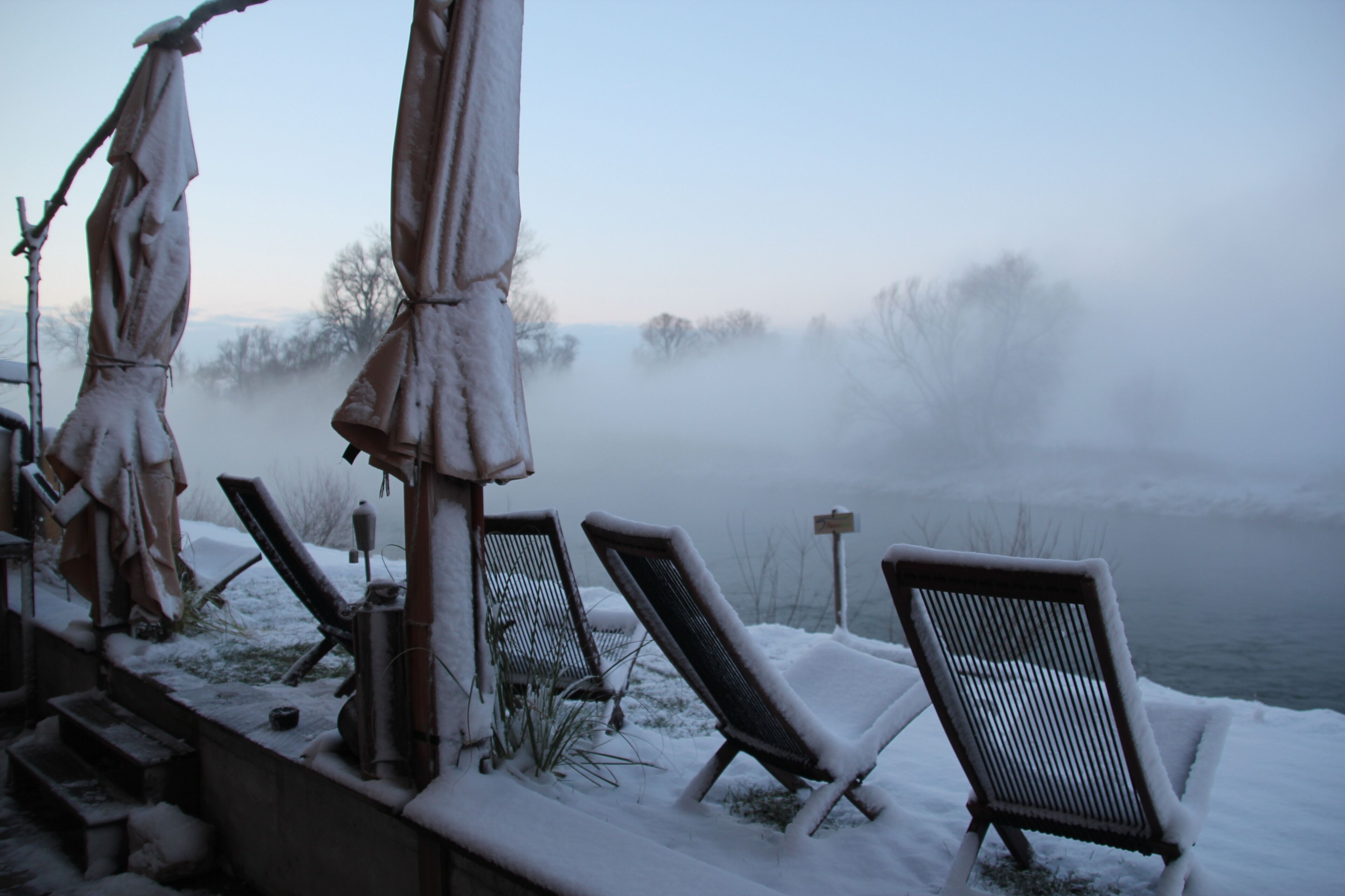A snow-covered outdoor seating area with lounge chairs and umbrellas, overlooking a foggy landscape.