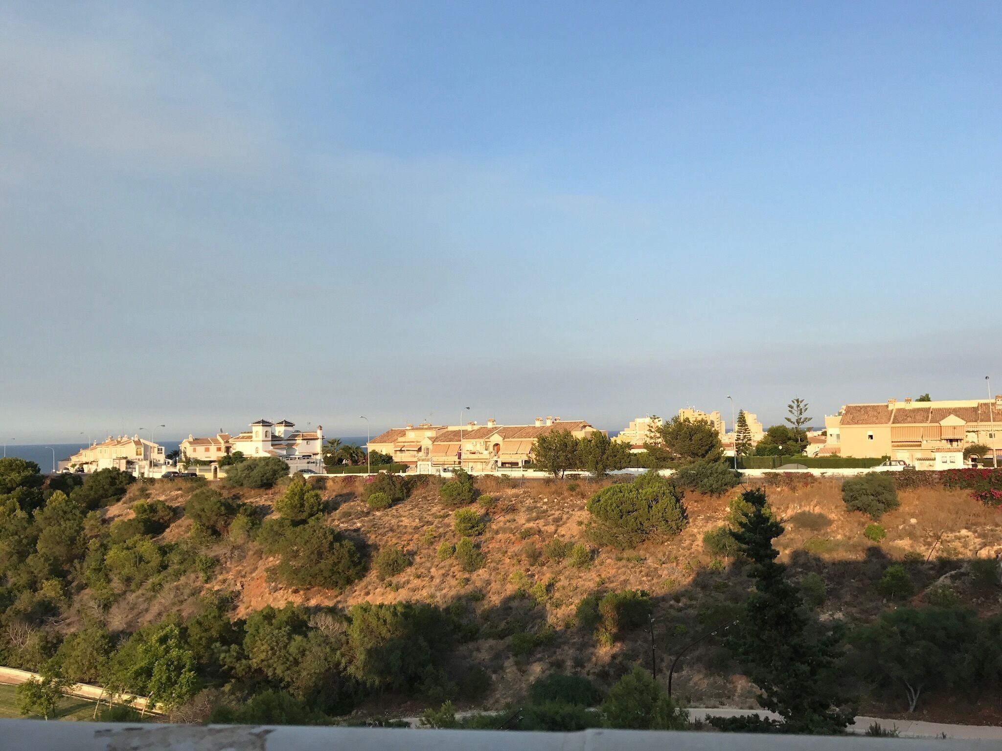 A coastal residential area with buildings on a hillside, trees, and a clear sky.