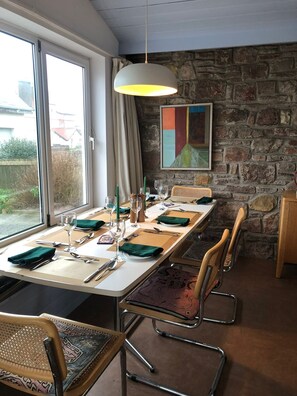 A dining area with a long table set for six, metal chairs, a hanging light fixture, a stone wall, and a window with curtains. - Bright modern holiday cottage steps from the beach and in the heart of village (Rhosneigr)