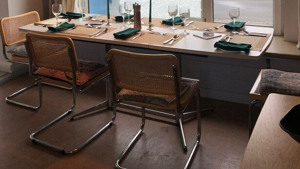 A dining area with a table set for four, a view of the sea through a large window, and a hanging light fixture. - Bright modern holiday cottage steps from the beach and in the heart of village (Rhosneigr)
